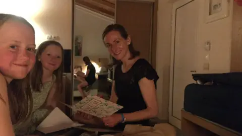 Jenny Sykes A woman is sat on a bed, holding a card that reads "mum" on the front. She is sat with two girls. All three are smiling whilst posing for the photograph. 