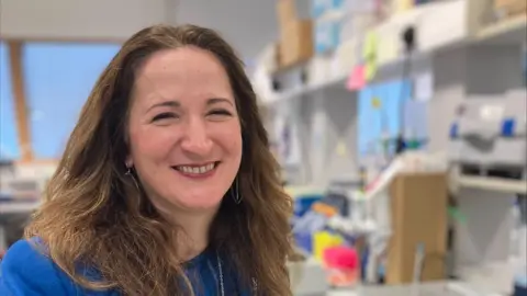 Professor Mirela Delibegovic is smiling at the camera. She is in the lab. She is wearing blue top and has brown shoulder length hair.