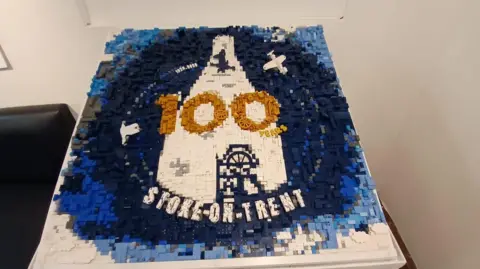 A Lego model of Stoke-on-Trent's centenary logo, which is made up of hundreds of grey, black, blue and white plastic bricks. Models of a Spitfire plane and a bottle oven can be seen in the middle, alongside the number 100 in gold Lego pieces and the word "Stoke-on-Trent" written in white along the bottom.