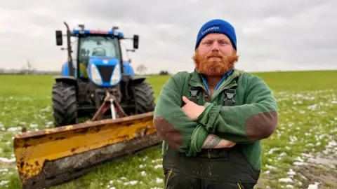 Sam Read/BBC A man in a green jumper and blue had stands in a field with melting snow. Behind him over his right shoulder is a blue tractor with a snow plough attached.