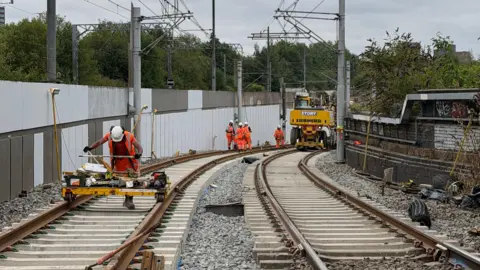 Five construction workers are on a tram track wearing orange high-vis jackets, one of them is pushing a yellow piece of machinery, the others are standing in the distance next to a vehicle.