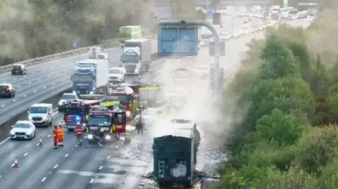 Fire engines, a smouldering lorry and long queues of traffic on a motorway. 