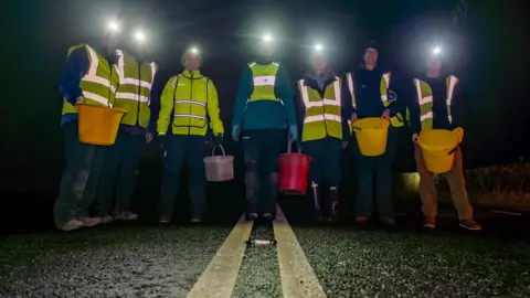 West Cumbria Rivers Trust A group of volunteers stand on a tarmac road. The volunteers wear fluorescent jackets and are holding brightly-coloured buckets which have frogs and toads inside. It is a dark night. 