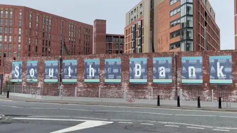 Sofía Luis-Hobbs/BBC A brick wall with the words 'South Bank' spelt out on blue boarded signs on it. Brick buildings are behind it.