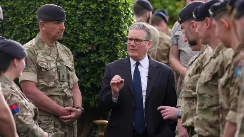 Sir Keir Starmer speaks to members of the military. He is stood in the centre with military personnel in uniform to his left and right. Sir Keir is wearing a dark-coloured suit, a white shirt and a dark-coloured tie.