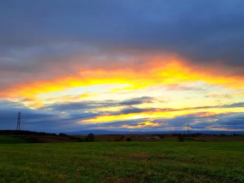 BBC Weather Watchers/Les at Large A view across fields with a string of pylons stretching into the distance, and a sky dominated by a setting sun shining through clouds