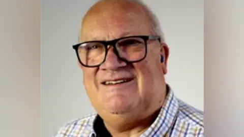 Rob Howard, a man wearing a checked shirt and glasses, is standing against a light-coloured backdrop.