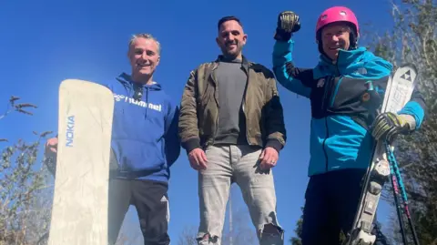 Three men, one dressed in ski clothes and one holding a snowboard, stand looking down at the camera from the top of an outdoor turf ski slope