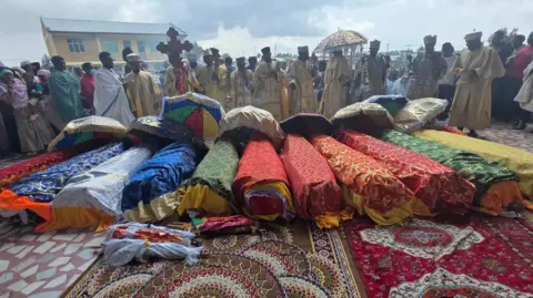 A row of brightly draped coffins laid before Orthodox clergy holding crosses and ceremonial umbrellas as grieving relatives and worshippers gather around