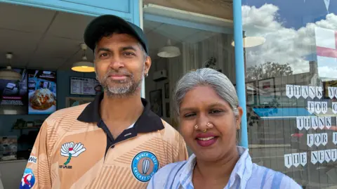 Michelle Lyons/BBC Rameswaran Selvarathinam and Sandhiya Prasad are pictured stood outside Munch Cafe. He is wearing a Munch Cafe cricket-style shirt, with the Yorkshire Cricket logo on, and a baseball cap. She is wearing a light blue shirt, has grey hair and dark pink lipstick. She is wearing nose rings.