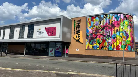The Way A large multi-coloured mural on the brick wall on a building, with lots of different patterned hands on it. There is a sign next to it saying "The Way".