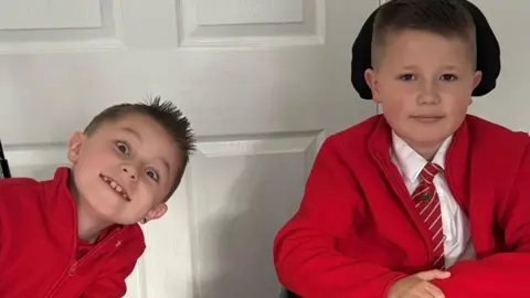 Two boys wearing red school uniforms smile at the camera while sitting side by side in front of a white panelled door. The boy on the right is seated in an electric wheelchair with a joystick control visible, wearing a shirt and striped tie under his red cardigan. The boy on the left leans in towards his brother, wearing a red zip-up fleece.