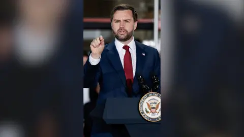 JD Vance in a navy suit with a red tie speaks at a podium. His arm is raised.