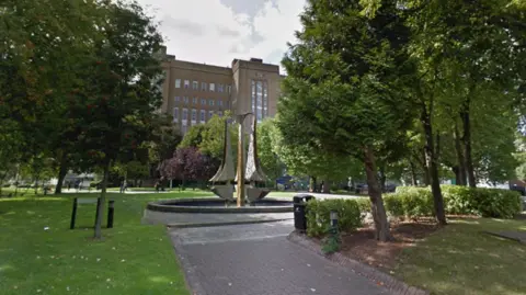 An early 20th-Century brick building, approximately six stories high, is flanked by trees and green space with a modern sculpture, Tipping Triangles, in a water feature in the foreground. The sculpture consists of three 18-foot stainless steel "triangles" with one curved side. 