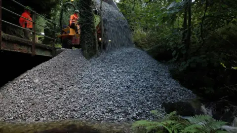 Large amounts of gravel are being poured by workmen next to a river. The pile of gravel is large. Two workmen, wearing orange hi-vis outfits, are standing on a platform next to the top of the gravel pile.