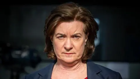 A head and shoulders framed picture of Eluned Morgan in artificial light, with a blurred and very dark background behind her. She has a very serious expression on her face.