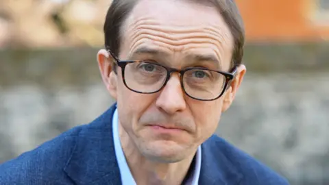 A close-up shot of BBC's political editor Chris Mason looking straight to the camera. He wears brown glasses, a blue shirt and blue blazer.