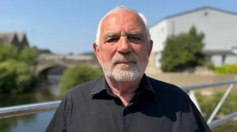 A man with grey hair and a grey beard in a black shirt stands on a bridge with a river, trees and a white building behind him. The background is blurred but there looks like another bridge over the water behind him.