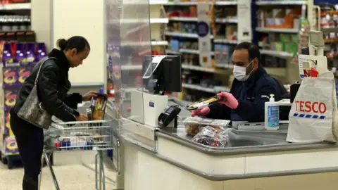 PA Media A Tesco supermarket cashier wearing protective face mask and gloves assists a shopper behind a plastic screen