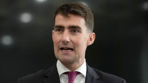 PA Media Jack Chambers is wearing a suit, white shirt and pink tie. He has short black hair. The background is blurry. 