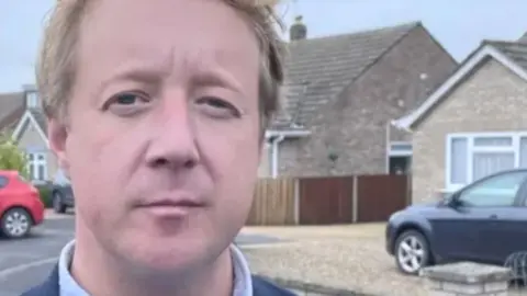 A head and shoulders image of Paul Bristow standing on a residential road, with bungalows in the background, a black car parked in a driveway and red car parked by a kerb.