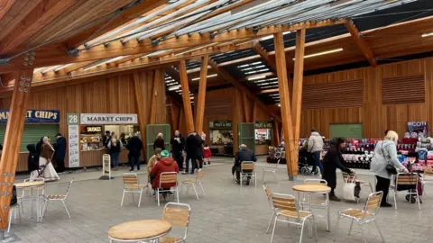 Great Yarmouth's market hall, a wooden structure with zinc roof. In the foreground is seating and tables, with people milling about inside the market building. Stalls can be seen in the backdrop of the building.