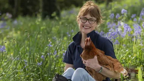 Simon Moore Jane Howorth, holding a hen in her right arm, sitting on the ground, wearing a dark blue top, and light blue trousers. She has on glasses and her hair is up. There are bluebells all around her in a field. She is smiling and looking straight at the camera. 