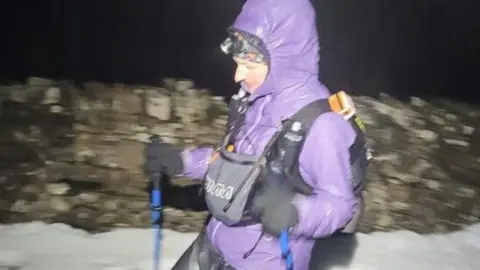 Mel Sykes A woman wearing a purple hooded coat and rucksack treks across a snowy terrain with walking poles. A dry stone wall is in the background and it is dark.