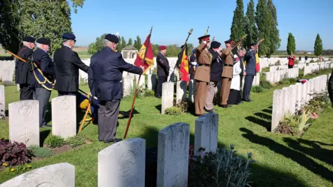 Crown Copyright Veterans standing at a row of graves. They are holding red, black and yellow flags.