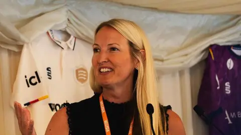 Rachel Lewis speaking on stage in front of a microphone with her right hand held out. She has long blond hair and a sleeveless black top