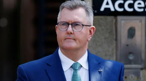 A man with short, cropped grey hair looks to the left of the camera. He has black-rimmed glasses and is wearing a blue suit, white shirt and green patterned tied. He is standing outside a building, a gateway can be seen, out of focus, behind him