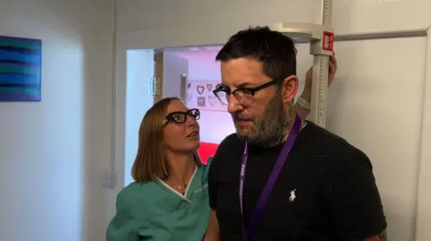 A man stood by a stadiometer, which measures height. He has dark hair and glasses on and is wearing a black T-shirt. There is a nurse in a green NHS uniform. She has glasses and is looking up, measuring the man's height.