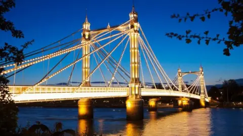 Albert Bridge is lit up at dusk with light bulbs across the spans of the structure and a turquoise sky behind. Lights are reflected onto the River Thames below.