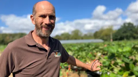 Paul Keene is smiling at the camera. He has a bald head with a grey beard. He is stood in a green field. He is wearing a brown polo top. 