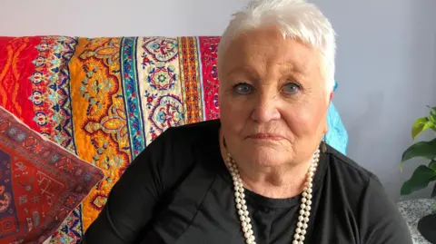 Pat Vinycomb looks at the camera. She has short, white hair and is wearing a black top with a string of white beads around her neck. Behind her on the chair is a colourful, patterned throw and cushions.