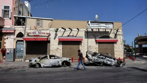 Reuters A man walks past two cars burnt during unrest in the Israeli city of Lod