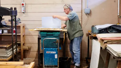 Carmel King Jim Patterson wearing blue jeans and a green t-shirt. He is standing in his workshop, lifting a large sheet of paper off a pile of crinkled handmade sheets.