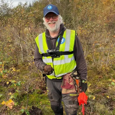 Prof Tony Pollard Ian Deveney is standing in a wooded area. He has a blue cap, glasses, a grey beard and is wearing a high visibility vest.