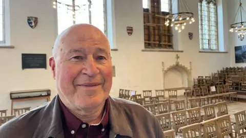 Andrew Turner/BBC A man standing inside a church, where the window to the right of him is boarded up. He is clean shaven, bald and wearing a burgundy shirt, with a grey/brown jacket over it. There are chairs placed and stacked behind him, and hanging chandeliers, which are lit.