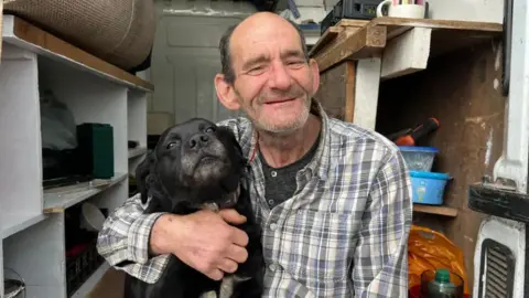 A man wearing a chequered shirt cuddling a black dog. He is sitting in the back of a van.