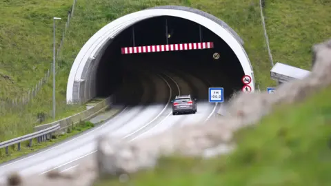 A grey car enters a tunnel, driving past red "no pedestrian" and "no cyclist" roadsigns. The tunnel disappears into a grassy hillside. A blue and white radio station information sign reads "FM 100.0".
