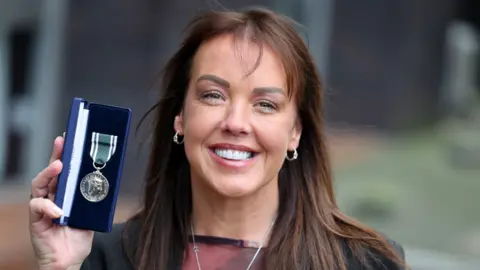 Andrea Raine has long brown hair with a long fringe. She is wearing silver hoops and a silver necklace. She is wearing a black blazer and a sheer red top. She is smiling while holding up a royal silver medal with a green ribbon, which is in a navy box.