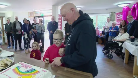 A room in Scott House is filled with dozens of people. In the foreground, young Oscar - who is wearing a red jumper and has his hair spiked up - is about to cut a cake which reads: "Happy Birthday", with his dad. Pink balloons in the shape of "10" are floating in the background.