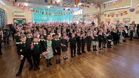 Children stood in a school hall waving Union flags