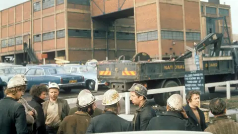 PA Media Archive image of miners wearing hard hats standing in front of a car park, with Lofthouse Colliery in the background.