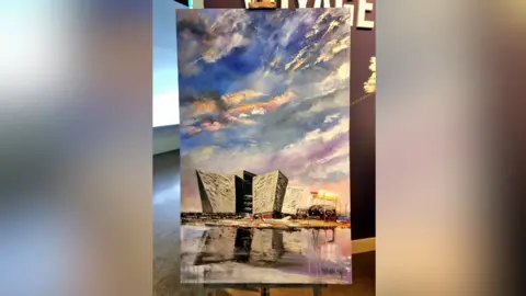 Stephen Whalley A portrait of the Titanic Building from the lough. The sun is behind the building. The picture is sitting on a canvas.