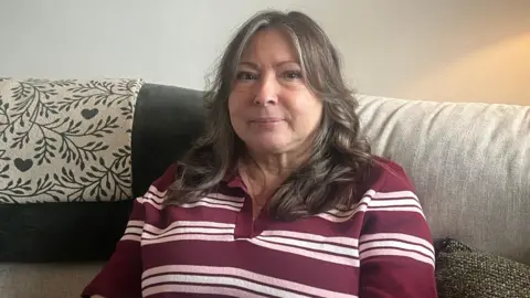 BBC A woman wearing a red striped sweater is sitting on a grey sofa. She has long dark hair and is looking directly at the camera.