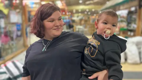 A young woman with cropped red hair and wearing a black top smiles at her baby son on her hip, who has a dummy in his mouth.