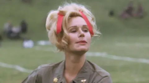 Screen Archives/Getty Images Sally Kellerman performs as "Hot Lips" Houlihan in army khakis with her blonde hair tied up with a red ribbon. She is looking off to the side and appears to be outdoors with grassy scenery behind her