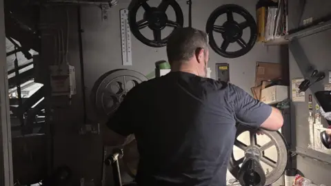 Ewan stands at a film cutting desk with his back to the camera. He is winding film onto a reel and concentrating on watching it go through smoothly. 
